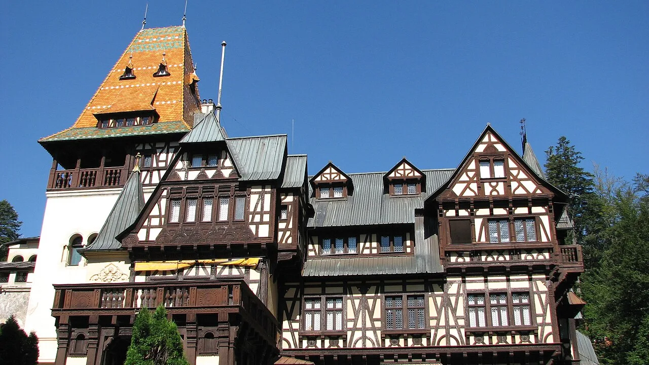 The half-timbered facade of Pelisor Castle in Sinaia under a deep blue sky, with its tower roofed in patterned orange and green sandstone tiles