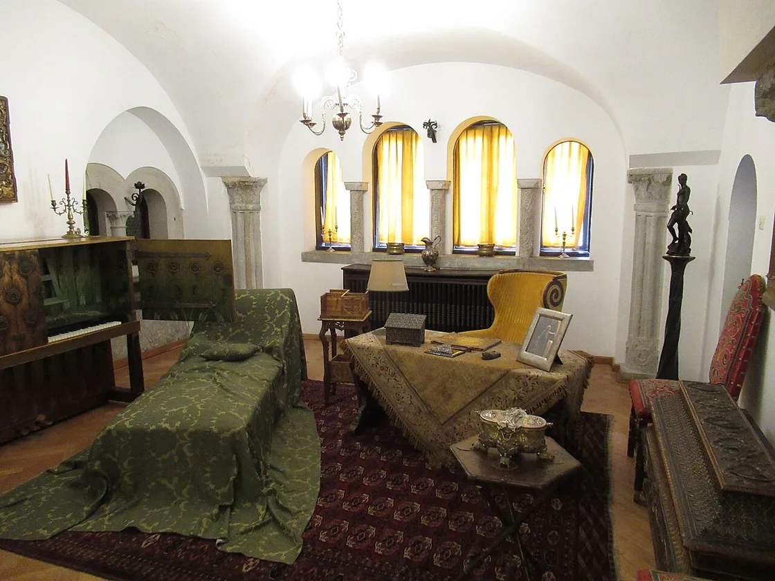 A vaulted room in Pelisor with carved stone columns, arched windows, a low divan under a green cover and oriental rugs on the floor