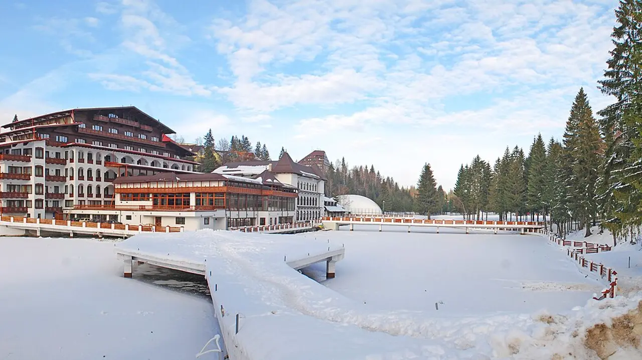 A large mountain hotel in Poiana Brasov under deep snow, a frozen pond and footbridge in front, pine forest and blue winter sky behind