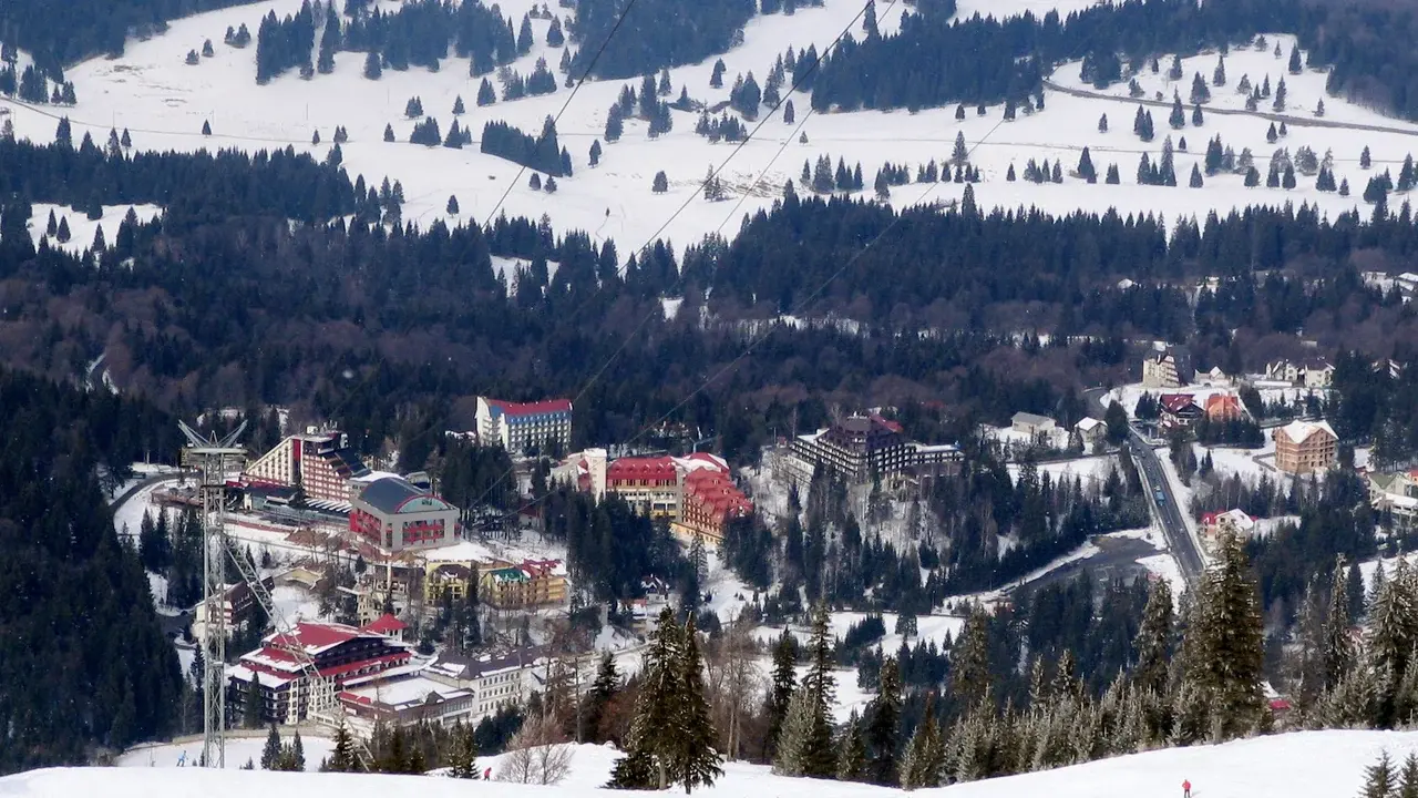 Poiana Brasov under snow seen from the ski slope above, with the cable car line crossing the frame and the access road curving in from the right