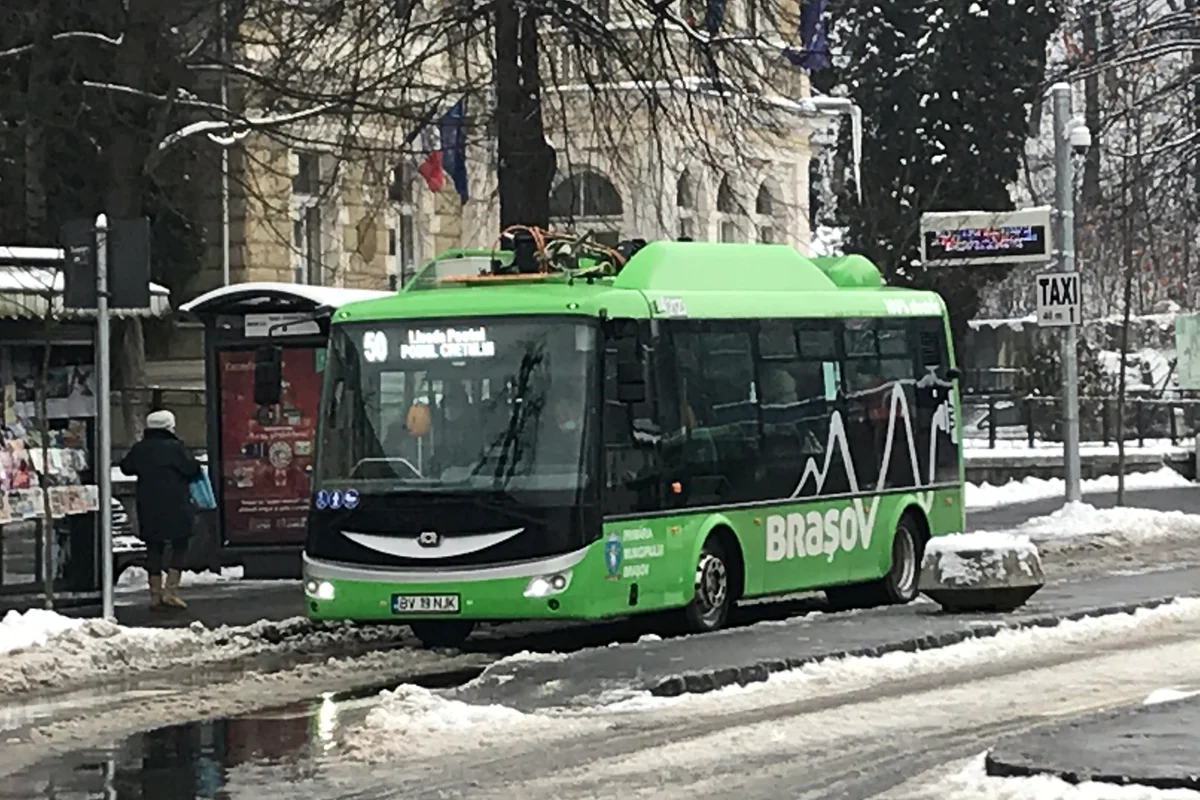 A green RATBV electric city bus standing at the Livada Postei terminal in Brasov in winter, snow banked along the kerb and a taxi rank sign behind