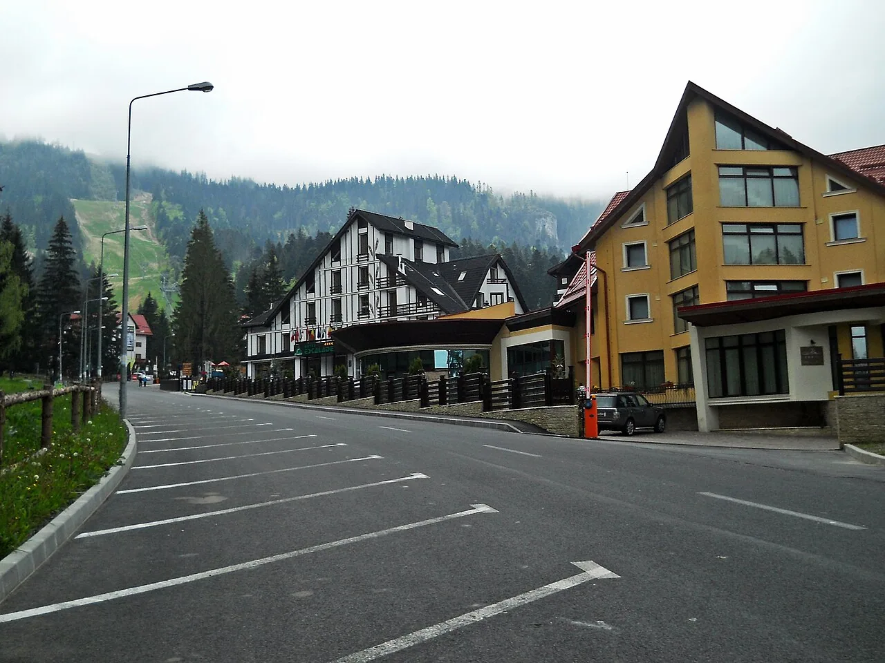 The main road through Poiana Brasov on a misty morning, empty marked parking bays along the kerb, hotels on the right and a ski slope disappearing into cloud on the left