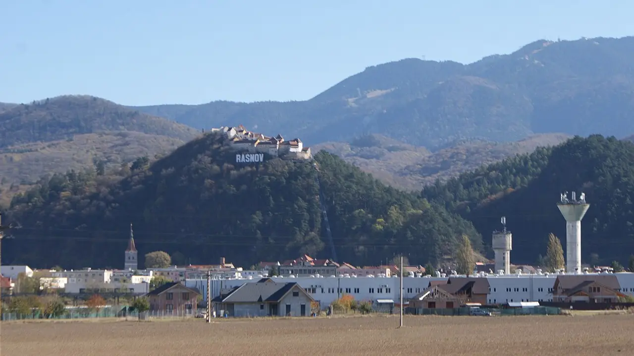 Rasnov Citadel on its wooded hill with the large white RASNOV sign on the slope below, the town at the base and the Barsa plain in front
