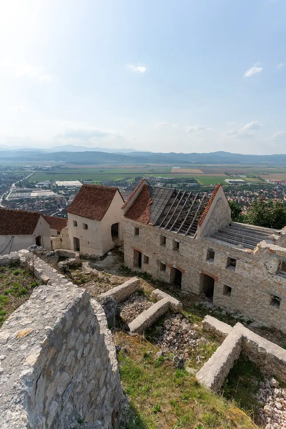 The ruined stone houses inside Rasnov Citadel with a sweeping view over the red roofs of Rasnov town and the flat green Barsa plain far below