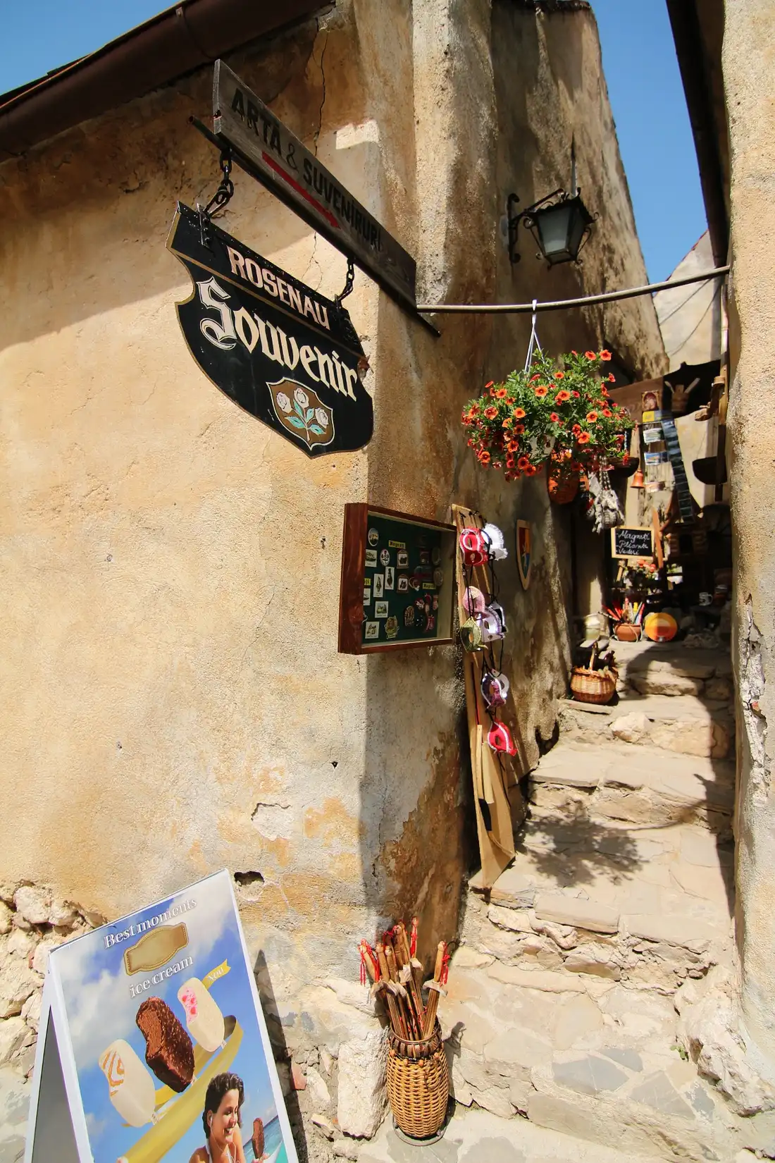 A narrow cobbled lane inside Rasnov Citadel, an old Rosenau souvenir sign and hanging flowers on a weathered plaster wall, souvenir stalls further down the alley