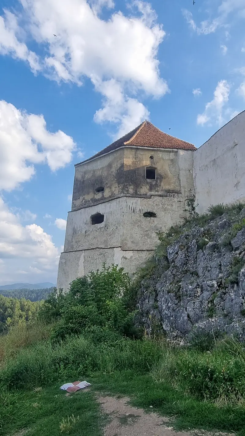A tall stone bastion tower of Rasnov Citadel with a red-tiled roof, rising above a limestone crag and green scrub under a blue sky with clouds