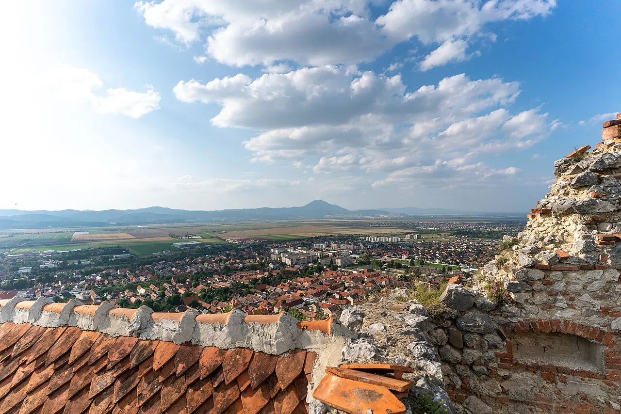 The panorama from Rasnov Citadel over the red rooftops of Rasnov town and the wide green Barsa plain, a lone mountain rising on the horizon under a bright sky