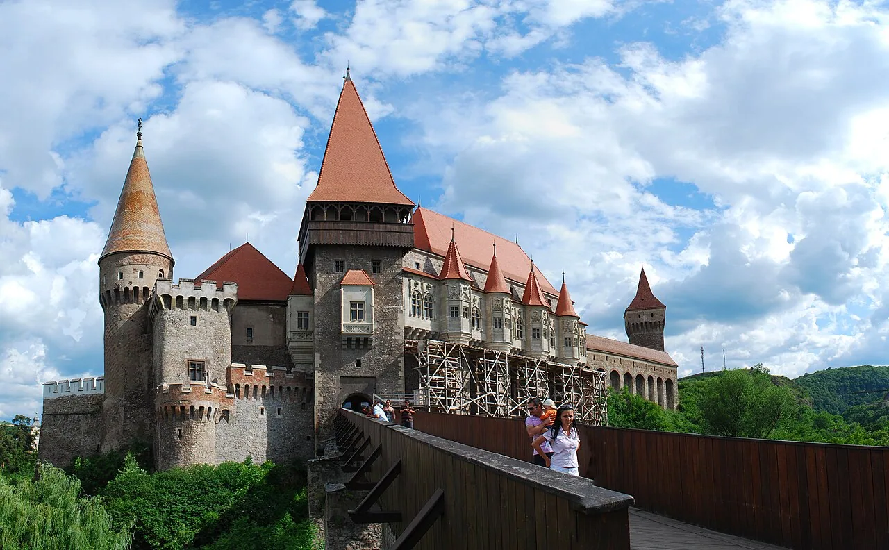 Corvin Castle in Hunedoara seen across its long wooden bridge, a mass of stone walls and tall towers with steep red roofs under a cloudy sky