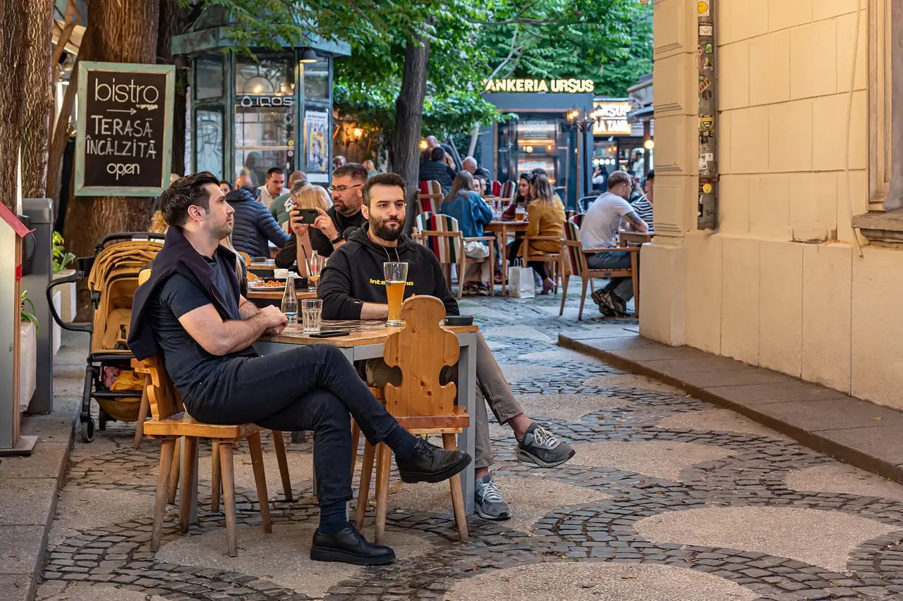 People sitting and working at an outdoor cafe terrace on a cobbled lane in Bucharest Old Town, one with a laptop bag, another checking a phone