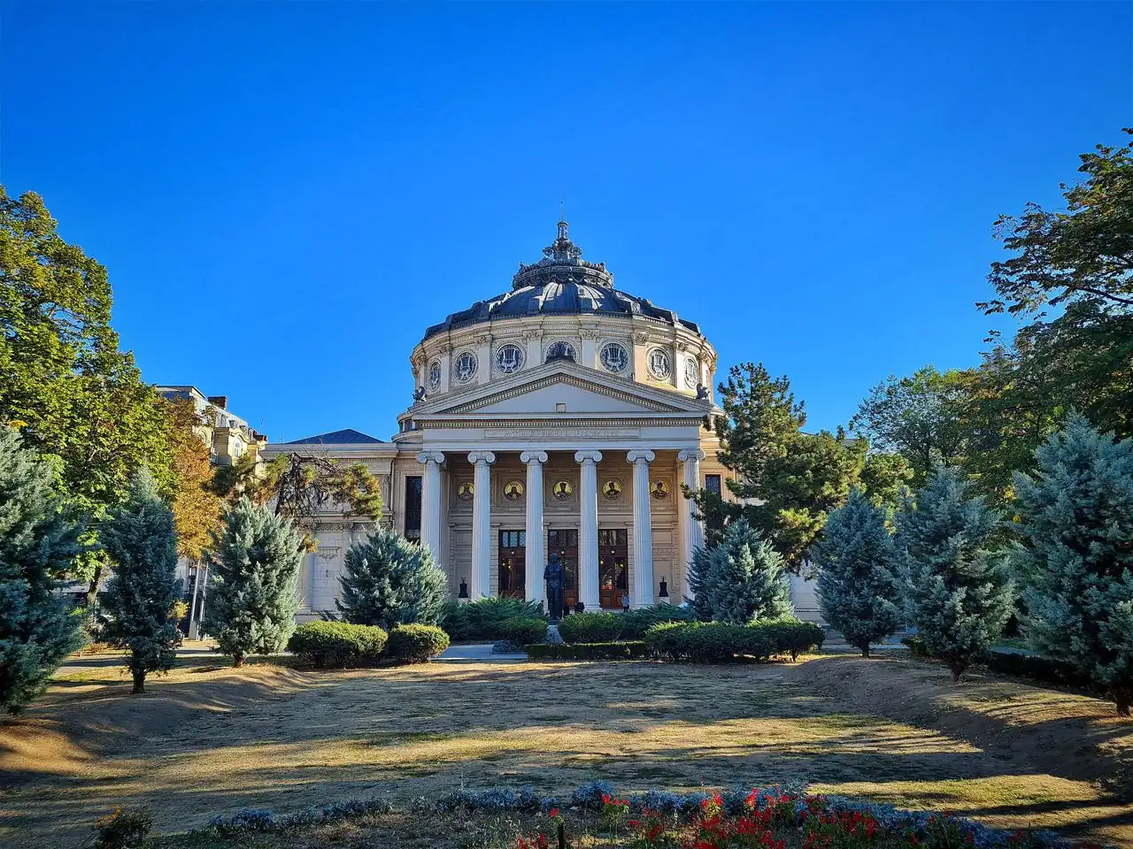 The Romanian Athenaeum in Bucharest, a domed neoclassical concert hall with an Ionic portico, seen across its garden on a clear autumn day