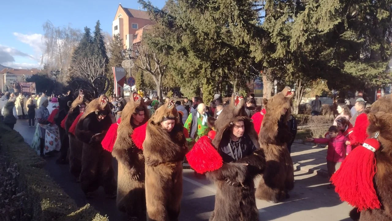 A line of dancers wearing whole brown bear skins with red tassels moving down a street lined with spectators in winter