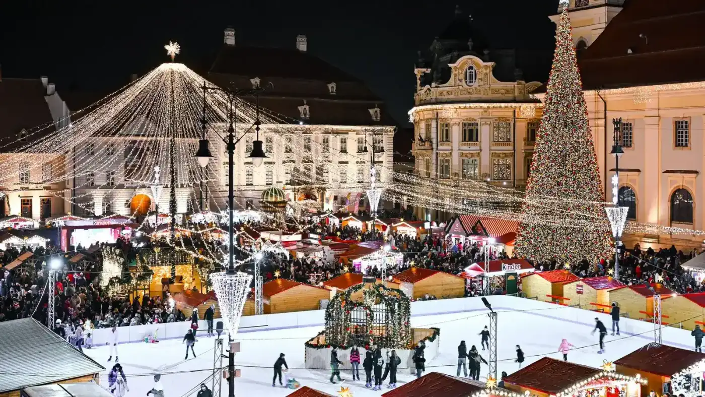Sibiu's Grand Square at night during the Christmas market, wooden chalets and a lit tree around a busy outdoor ice rink