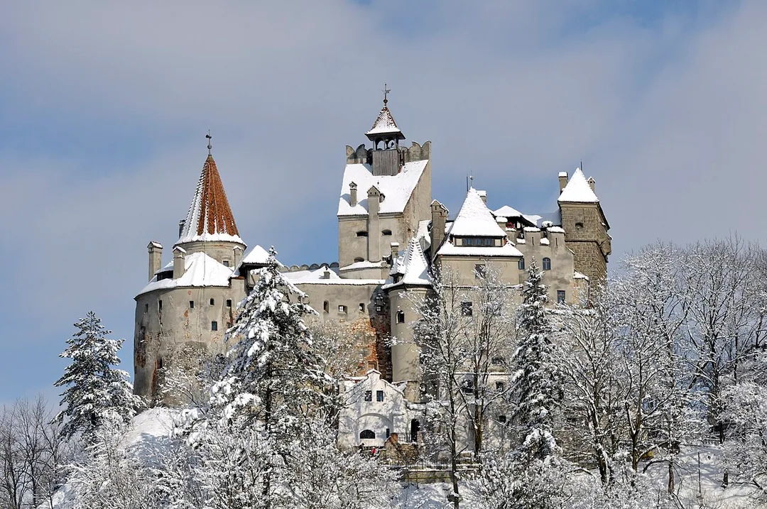 Bran Castle on its wooded hilltop under a heavy blanket of snow, bare and snow-laden trees framing the pale towers against a partly cloudy sky