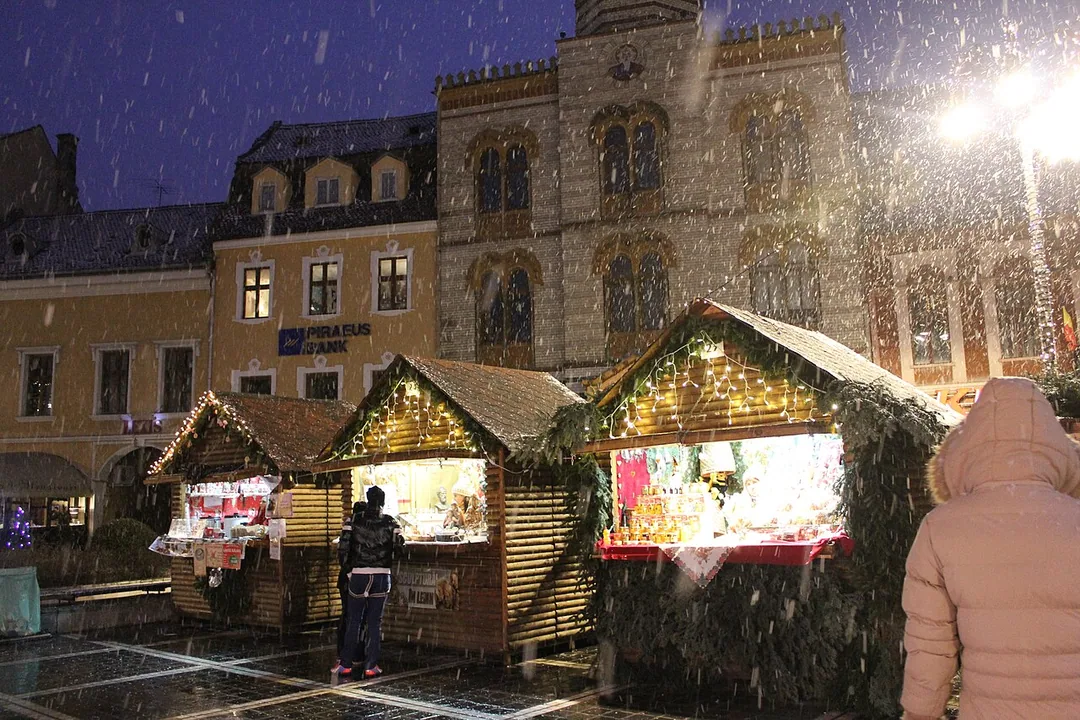 Snow falling at night over the wooden chalets of the Brasov Christmas market, lit stalls glowing against the old town facades behind