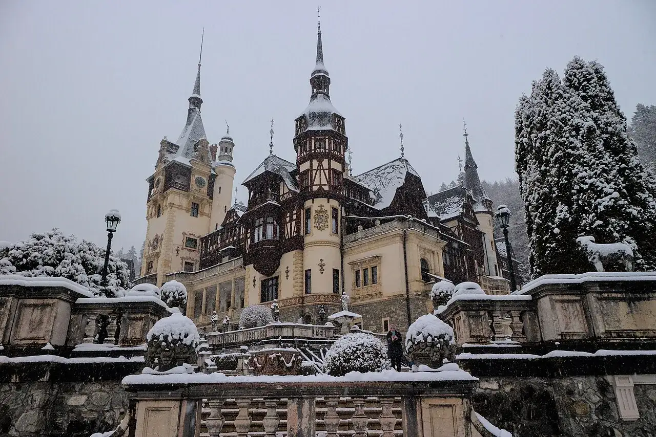 Peles Castle in Sinaia dusted in falling snow, its dark timber towers and pale stone facade rising above a snow-covered terrace and garden statues