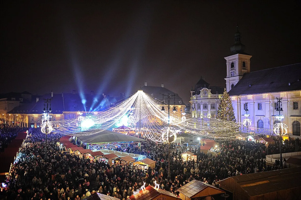 Sibiu's Grand Square lit up for the Christmas market at night, strings of lights fanning out over crowded wooden stalls and a lit Christmas tree