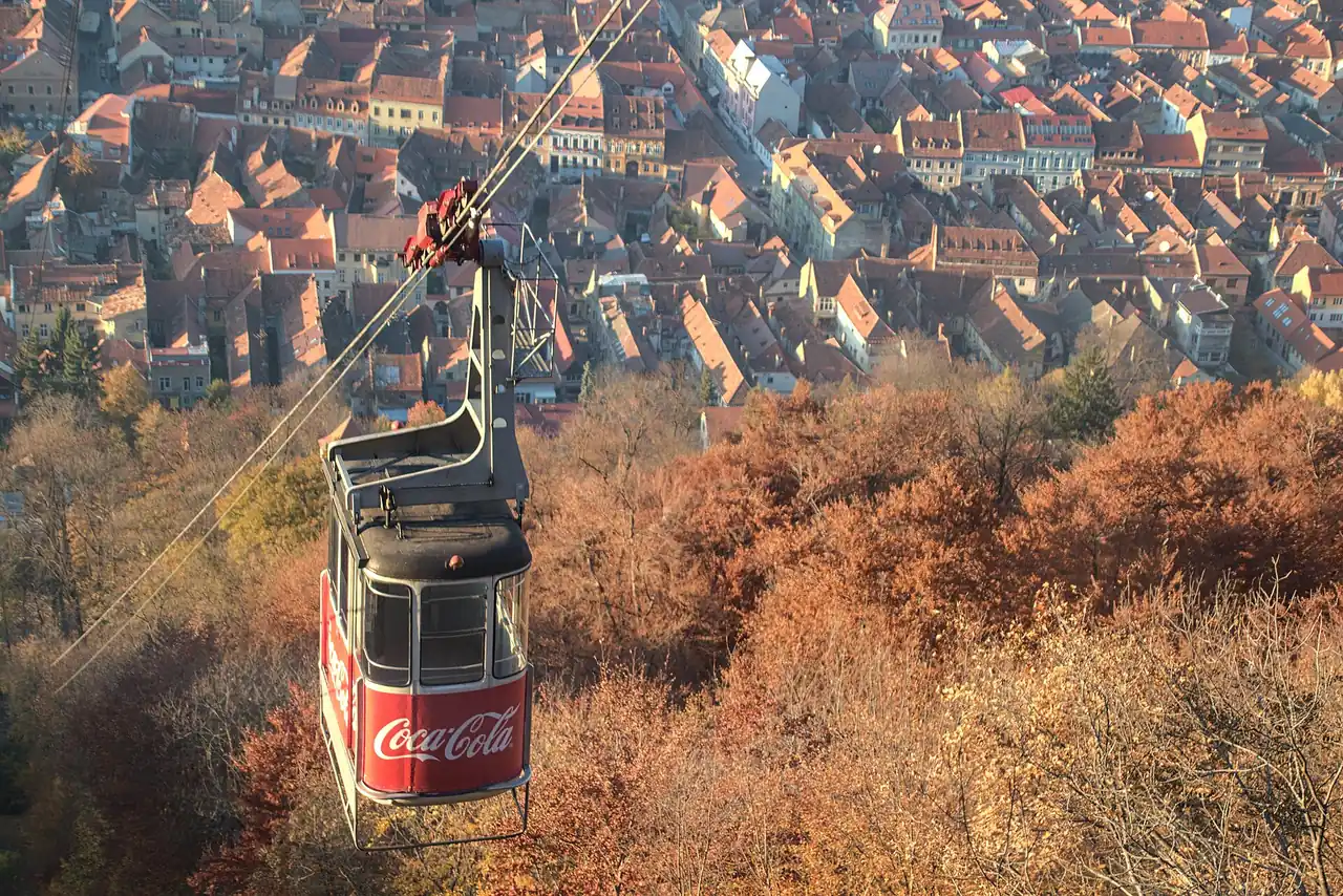 A red cable car cabin hanging above autumn woodland with the red rooftops of Brasov old town spread out below