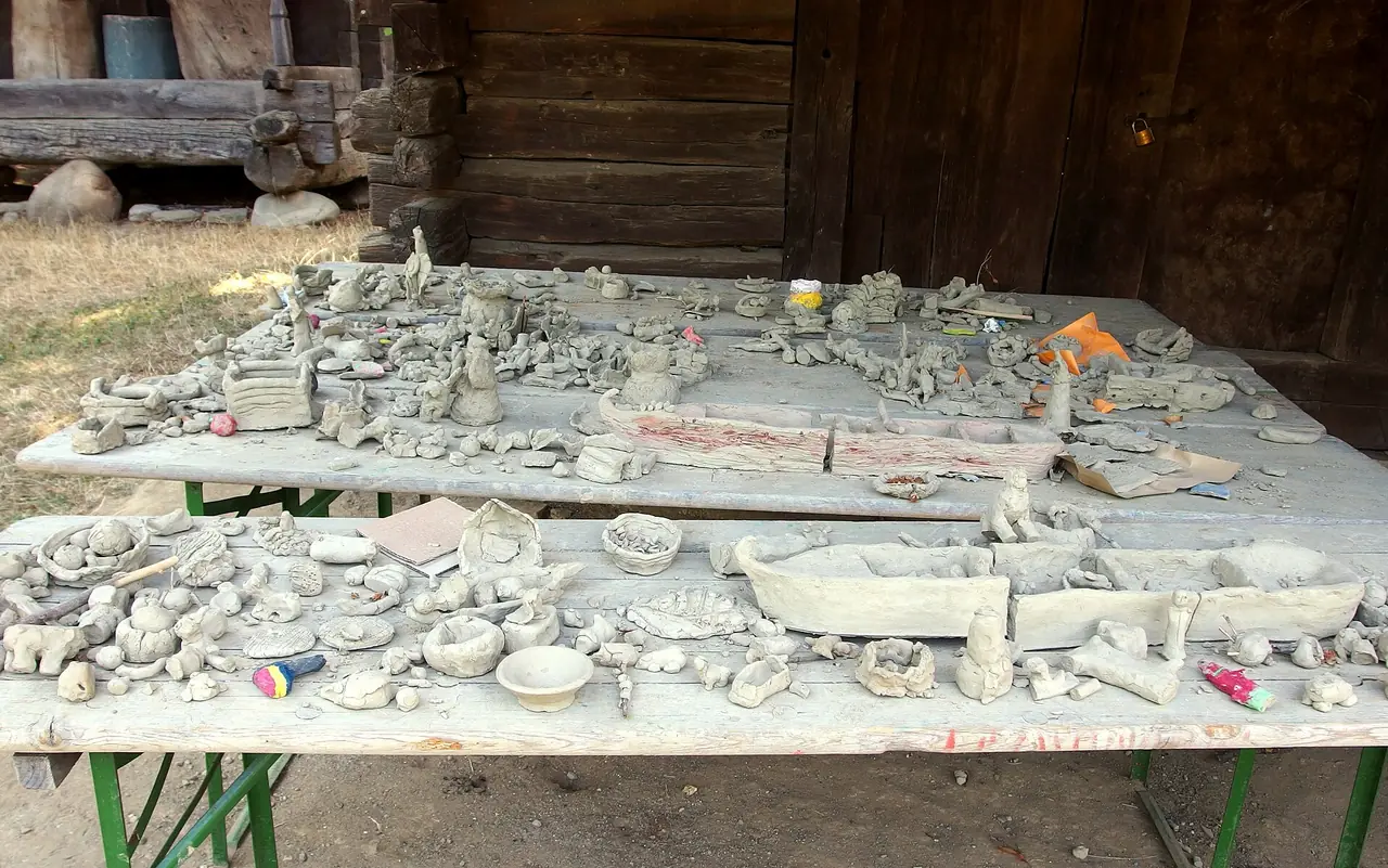 Dozens of small unfired clay figures and pots laid out to dry on wooden trestle tables outside a timber house at the Village Museum