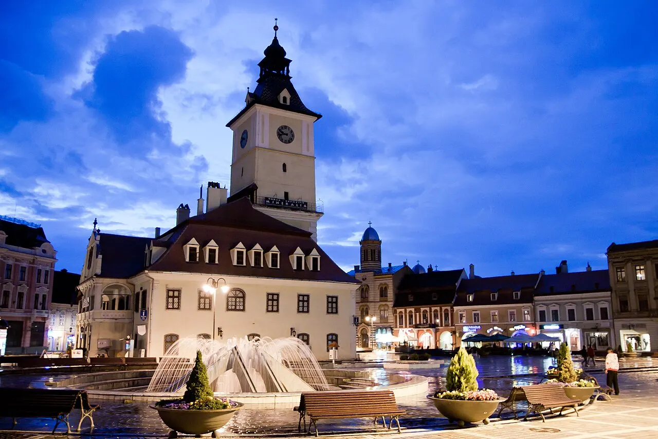 Council Square in Brasov, a wide cobbled plaza ringed by colourful merchant houses and the old town hall tower under a blue sky