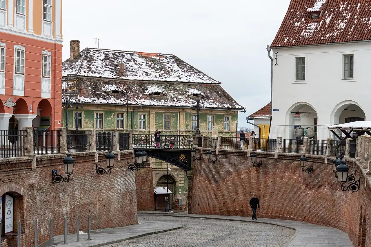 The wrought-iron Bridge of Lies in Sibiu over a lane in the Small Square, colourful old houses behind it under winter snow