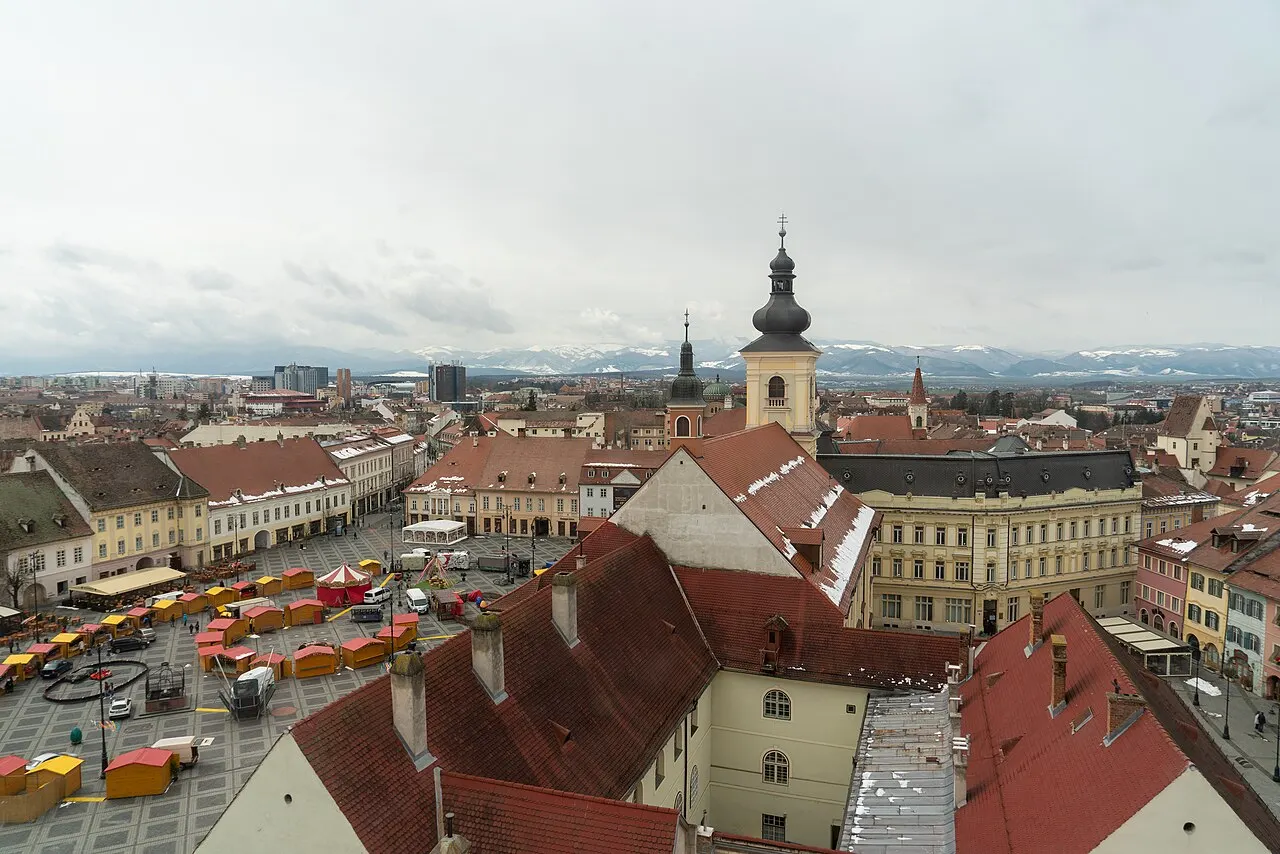The Large Square of Sibiu seen from above, a broad plaza framed by pastel baroque houses with the green dome of the Holy Trinity Church behind
