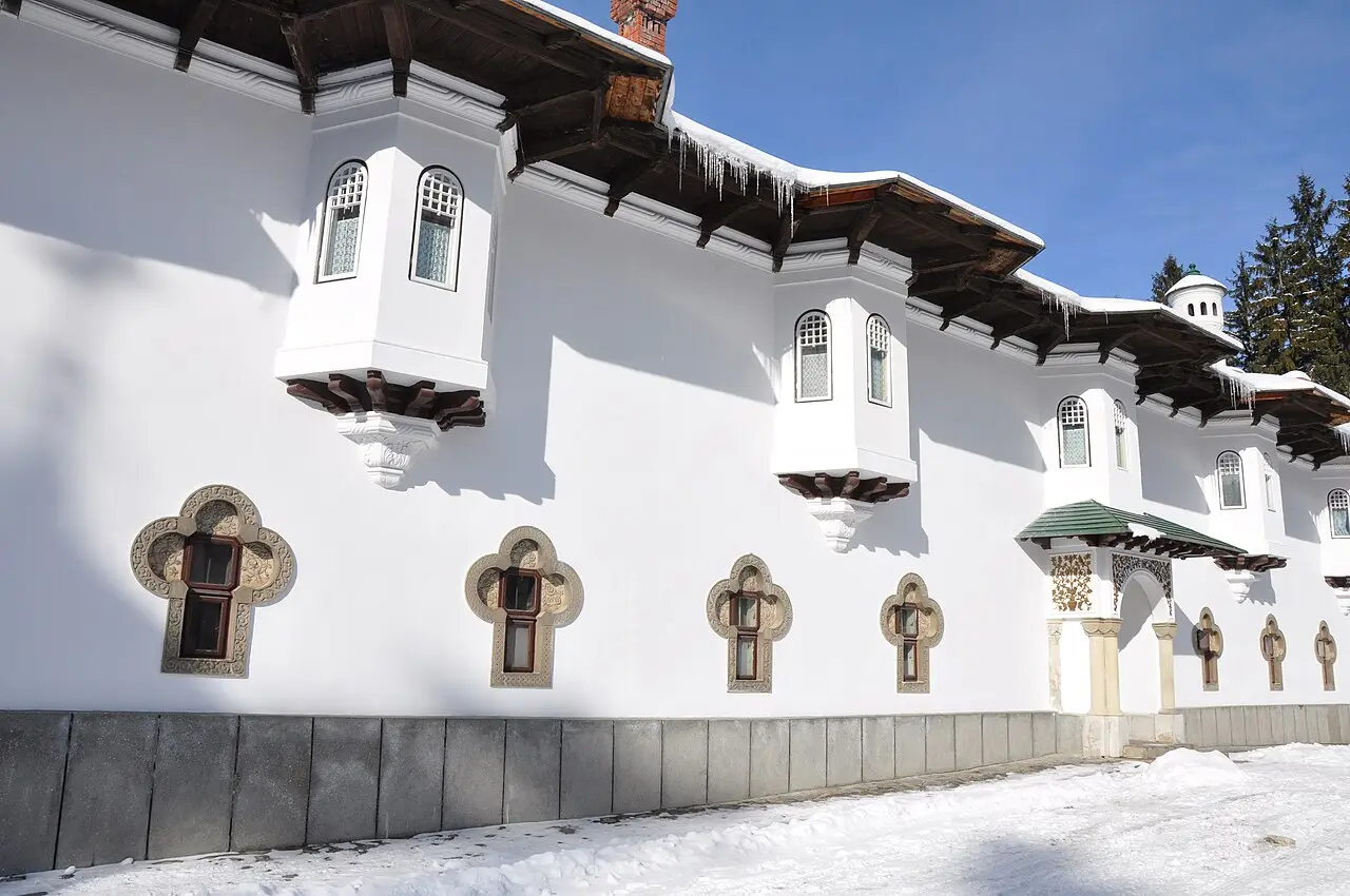 The whitewashed wall of Sinaia Monastery in snow, lined with ornate stone cross-shaped window frames and carved brackets under the eaves