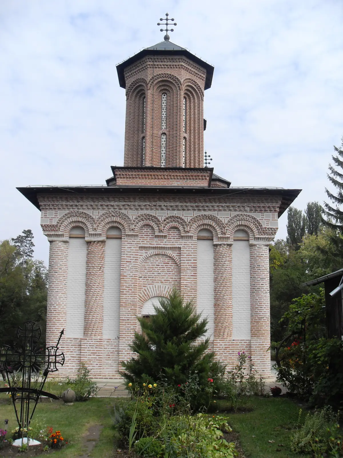 The striped brick apse of Snagov Monastery church with a small iron cross in the flower bed