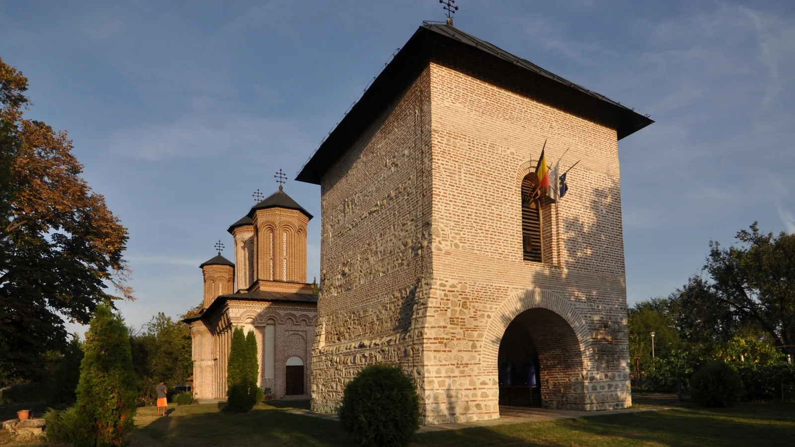 The brick church and stone bell tower of Snagov Monastery on a green lawn, with a visitor walking the grounds
