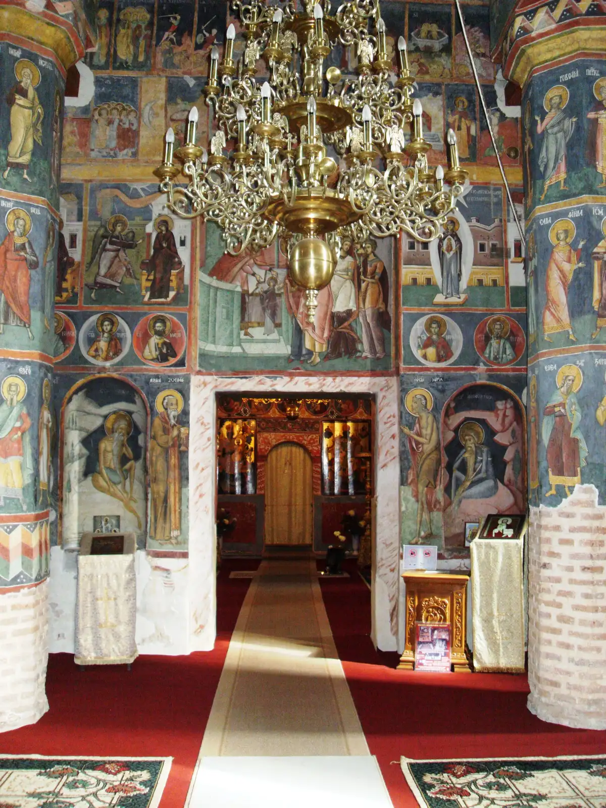 Frescoed nave of Snagov Monastery looking toward the altar, with painted columns, a brass chandelier and a red carpet