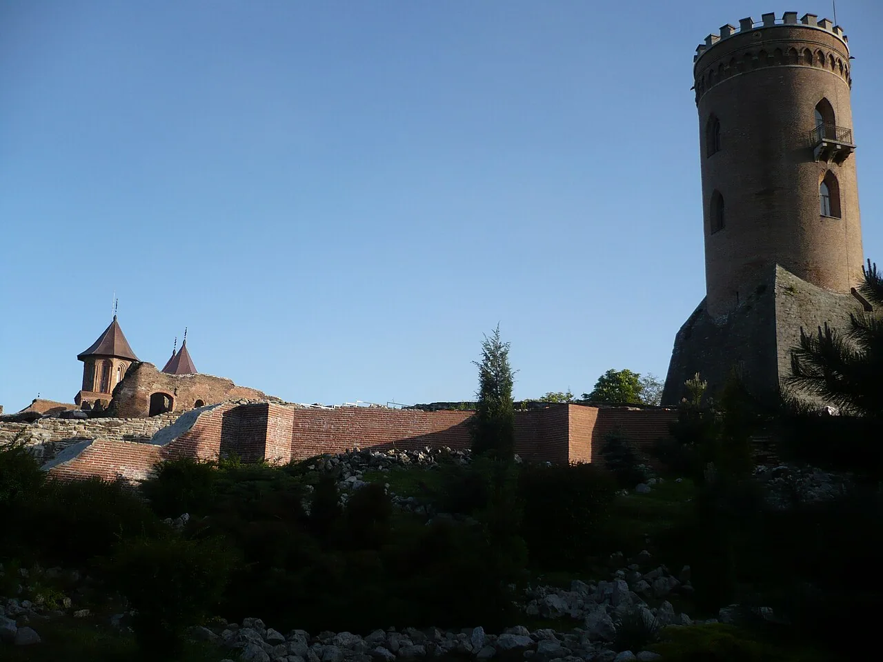 The brick Chindia Tower rising above the ruined brick walls of the Princely Court in Targoviste, with a small church visible beyond the ramparts