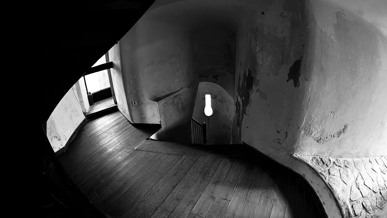 A black and white view down the spiral wooden staircase inside the Chindia Tower, worn plaster walls curving around a narrow stairwell