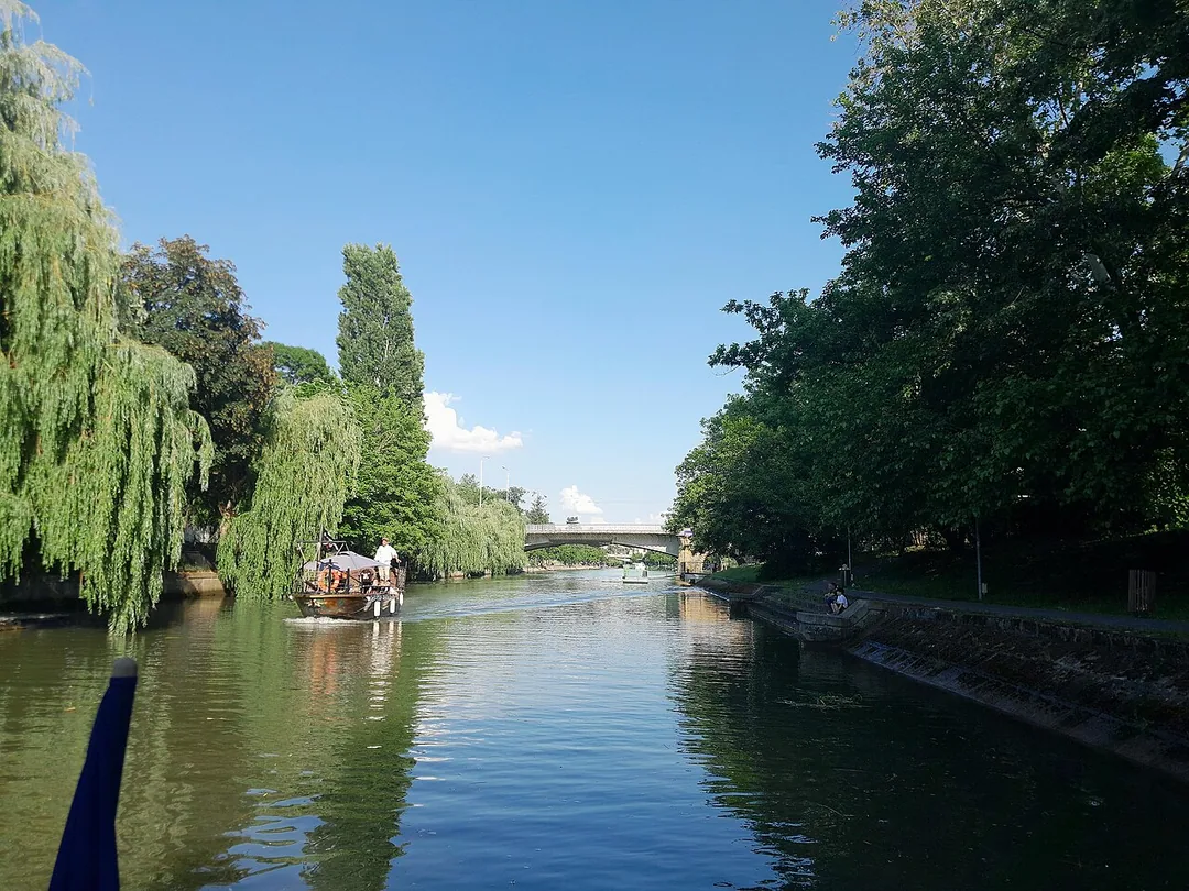 The Bega canal in Timisoara seen from a water bus, tree-lined banks and low bridges reflected in the calm green water