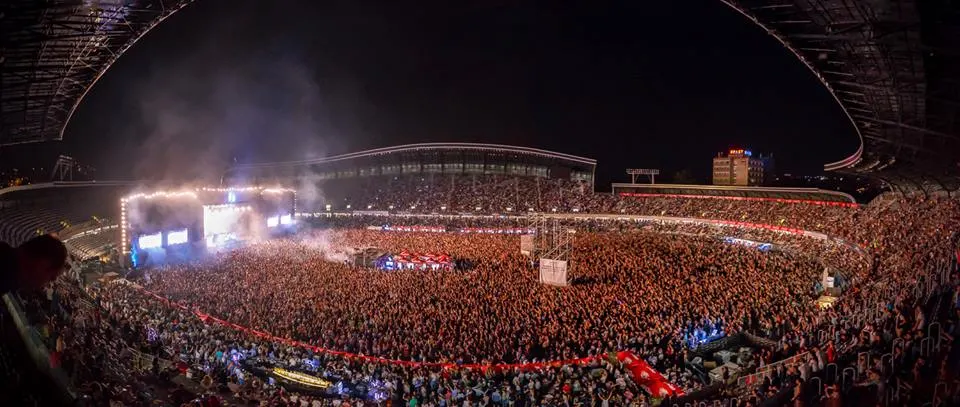 A packed crowd filling Cluj Arena at night during Untold, hands raised toward a brightly lit main stage with pyrotechnics and smoke