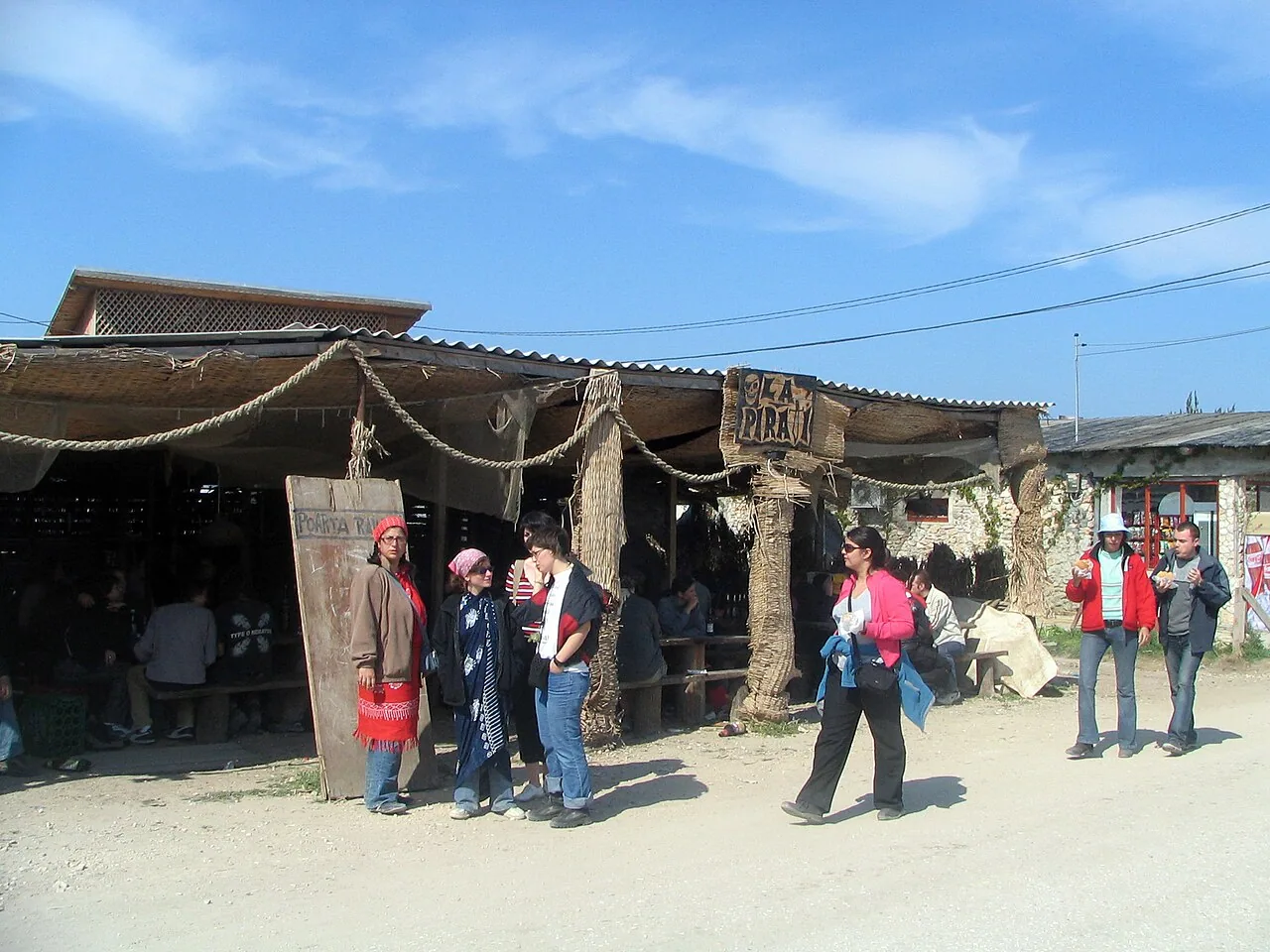 Visitors gathered outside La Pirati, a driftwood and thatch beach bar in Vama Veche with a hand-painted sign, on a sunny day