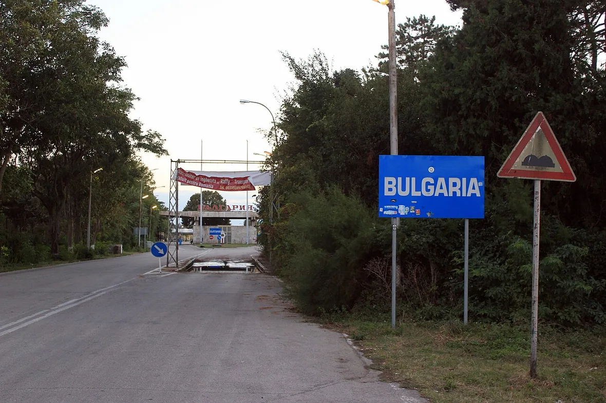 The road border crossing into Bulgaria at Vama Veche, a blue Bulgaria sign and checkpoint gantry visible down the tree-lined road