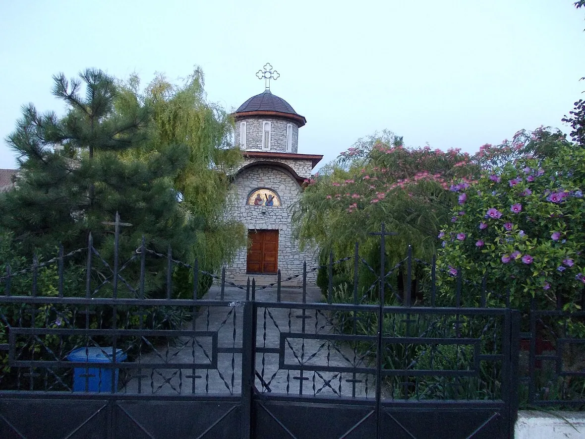 A small whitewashed Orthodox church with a domed bell tower in Vama Veche, framed by trees and flowering shrubs behind an iron gate