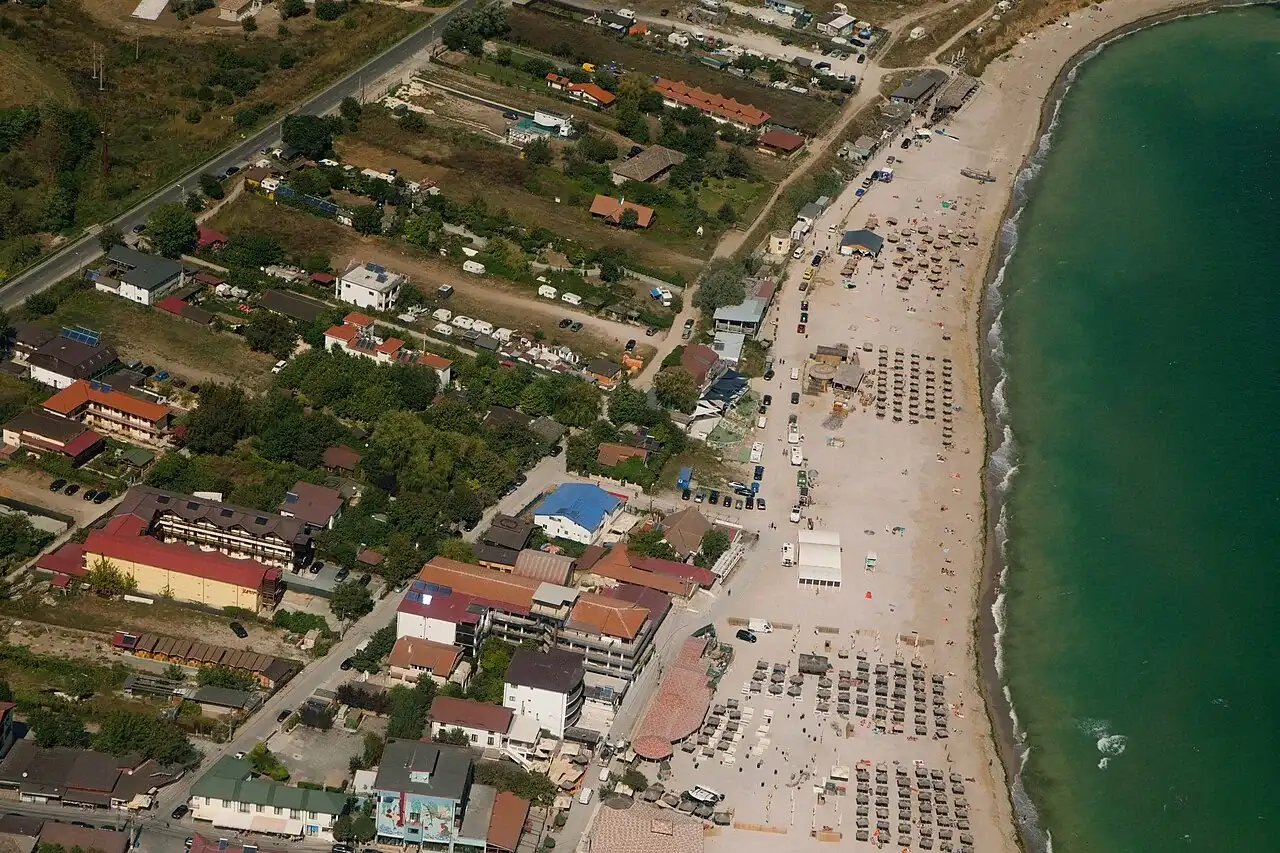 Aerial view of Vama Veche, low guesthouses packed along a curve of sandy beach against the green Black Sea