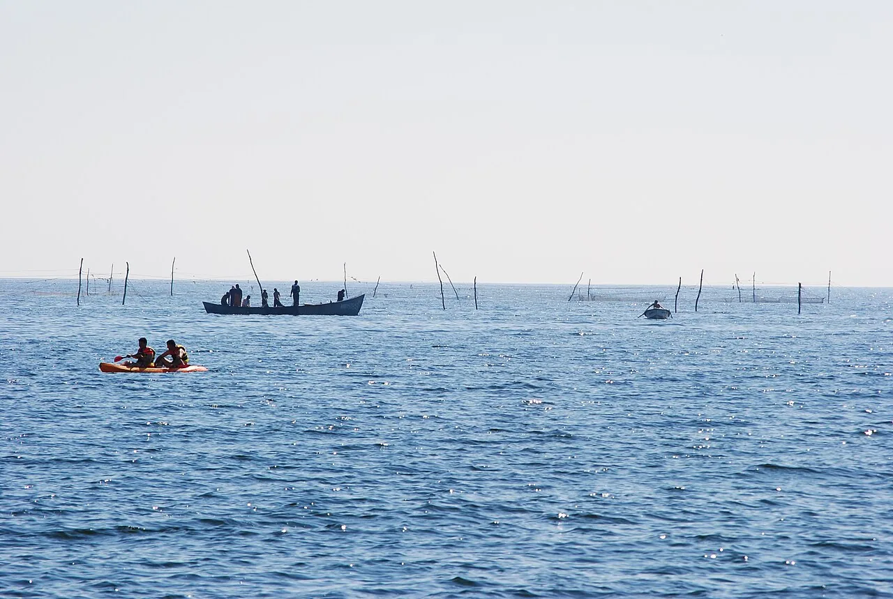 A traditional fishing boat with several fishermen and a kayak nearby in the calm waters off Vama Veche, fishing net markers visible on the horizon
