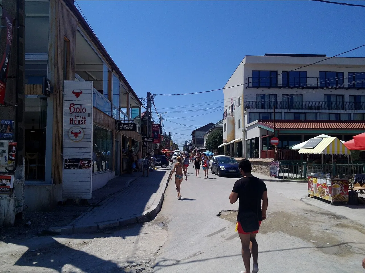 Looking up a sandy Vama Veche street from the beach toward guesthouses and parked cars under a bright sky