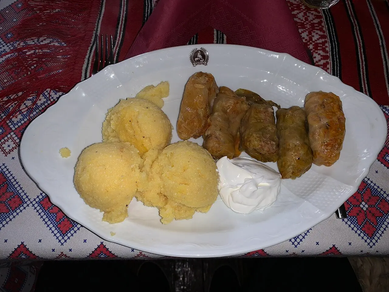 A plate of sarmale cabbage rolls with scoops of mamaliga polenta and sour cream on an embroidered Romanian tablecloth