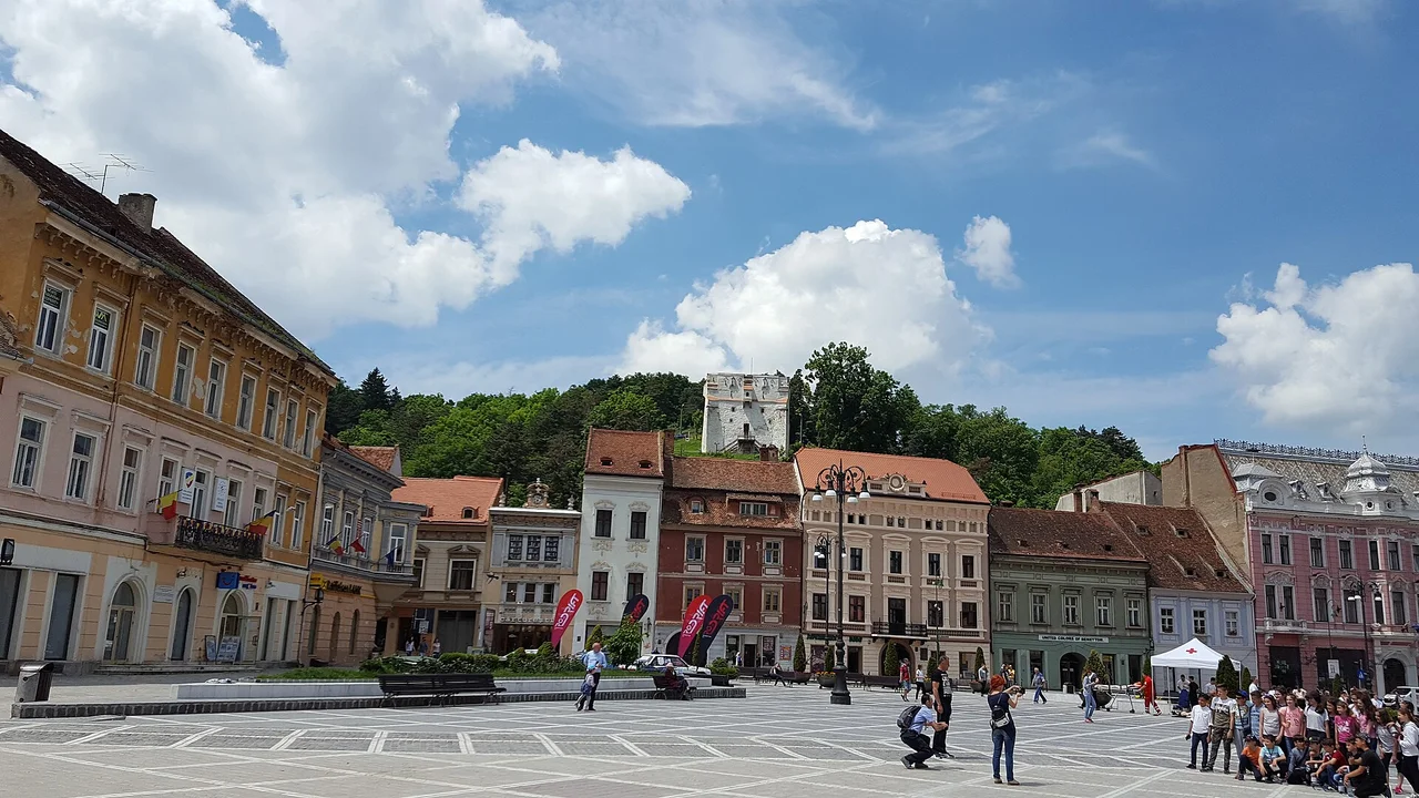 The colourful merchant houses of Council Square (Piata Sfatului) in Brasov old town, the White Tower on the wooded hill behind, on a bright summer day