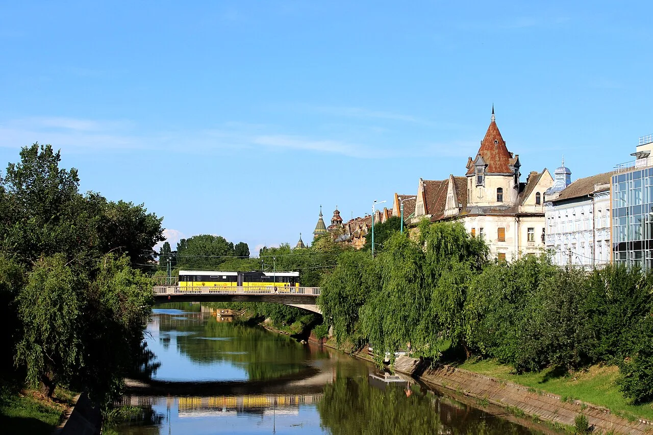 A yellow tram crossing a bridge over the Bega river in Timisoara, the turreted Palatul Ancora and riverside buildings reflected in the water under a blue sky
